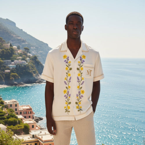 Man wearing a floral embroidered shirt and pants standing on a scenic coastal path.